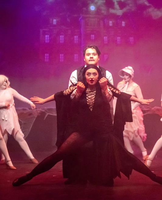 Morticia and Gomez Addams striking a dramatic dance pose, framed by a gothic stage backdrop and eerie ensemble members Morticia and Gomez Addams striking a dramatic dance pose, framed by a gothic stage backdrop and eerie ensemble members